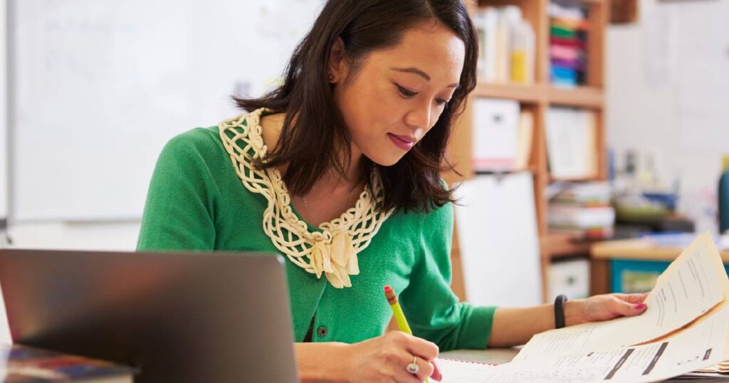 Female Asian teacher at her desk lesson planning and marking students work