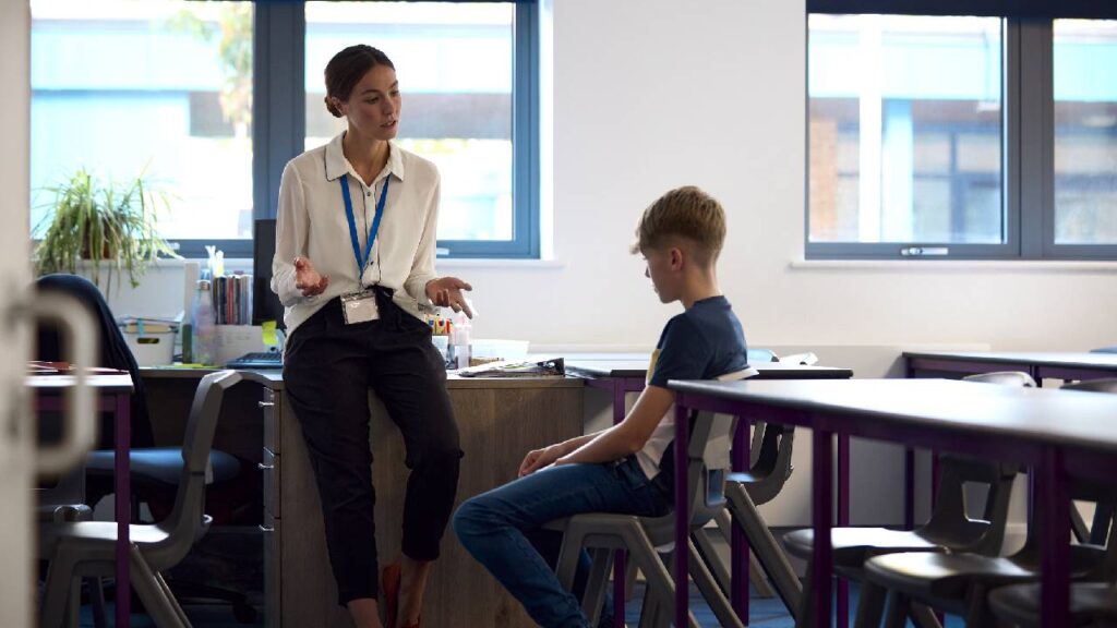 Secondary Or High School Teacher Sitting In Classroom With Unhappy Male Student After Lesson