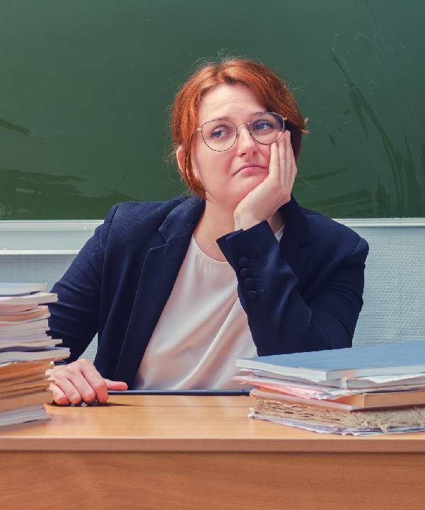 White female teacher who is sat at her desk. She is looking sad to be back at the school after the festive period.