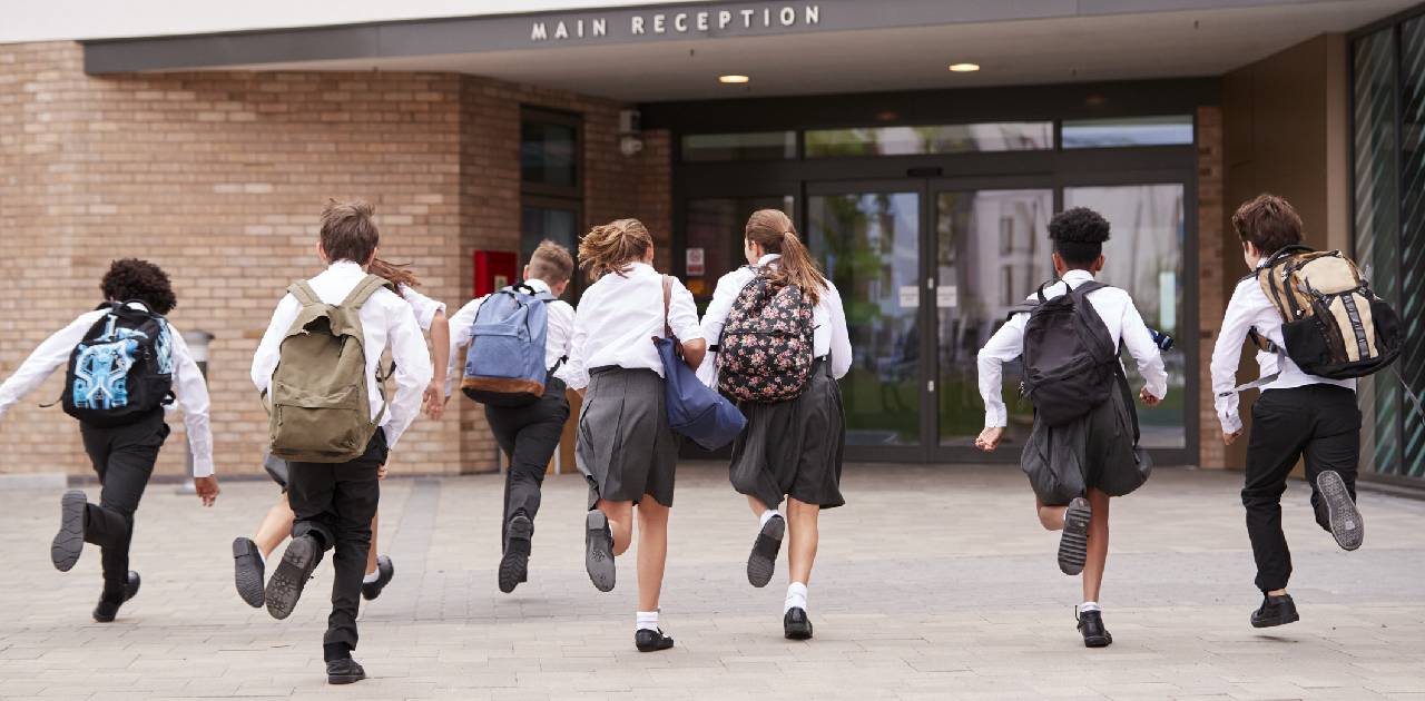 Group Of High School Students Wearing Uniform Running Into School Building At Beginning Of Class